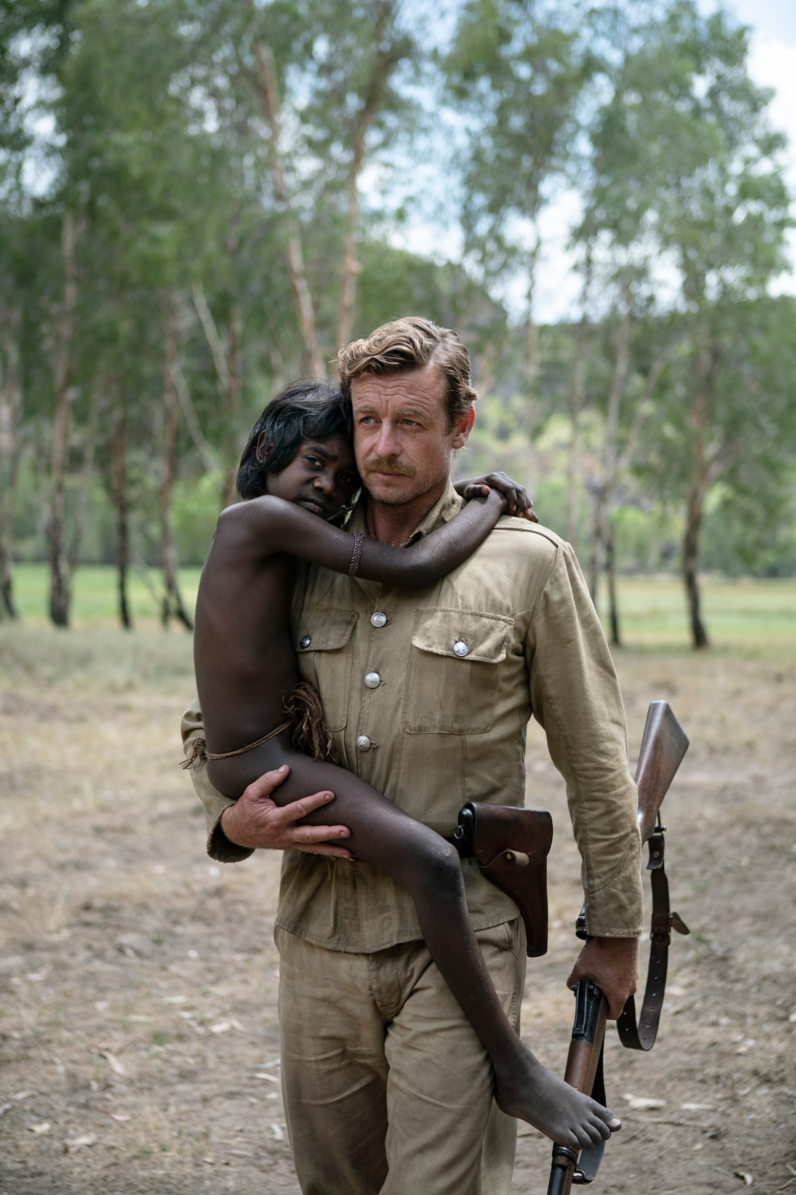 A white man in a uniform and holding a rifle is carrying a young Aboriginal boy in an outback setting.