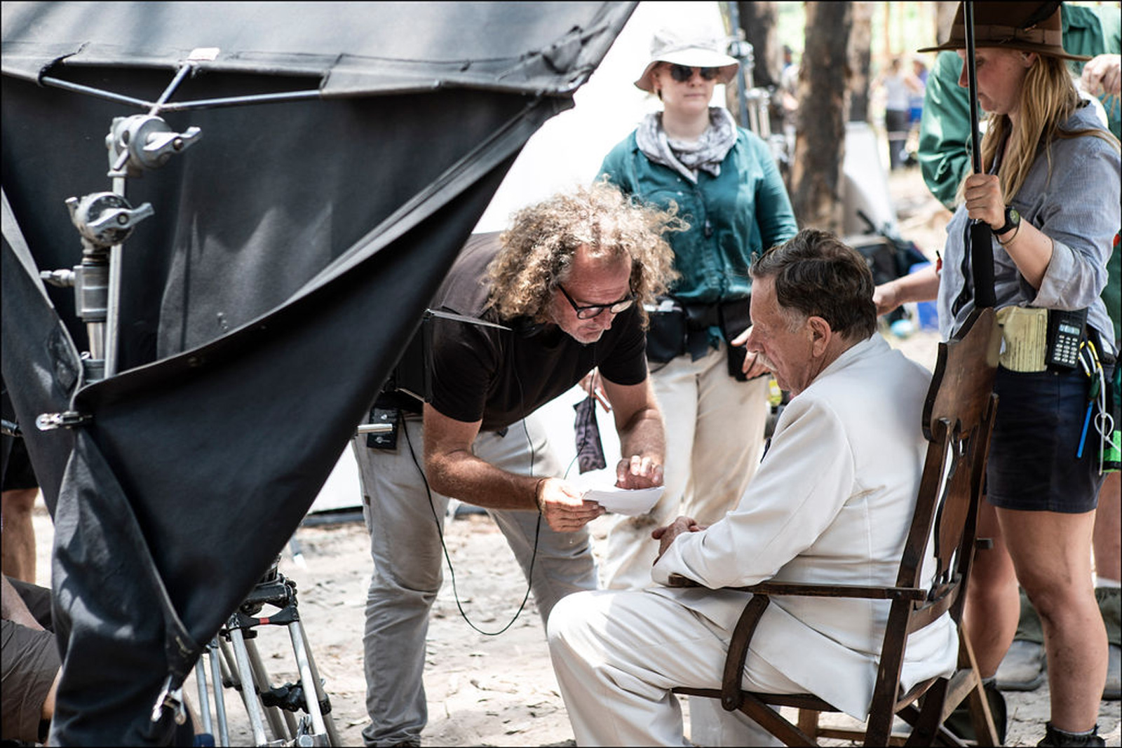 A behind the scenes image from a film production. A man sitting in a wooden chair in a hot, dry outdoor setting. He