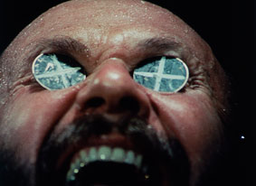Close up of a mans face, mouth open, with two-up coins with crosses on them in his eye sockets