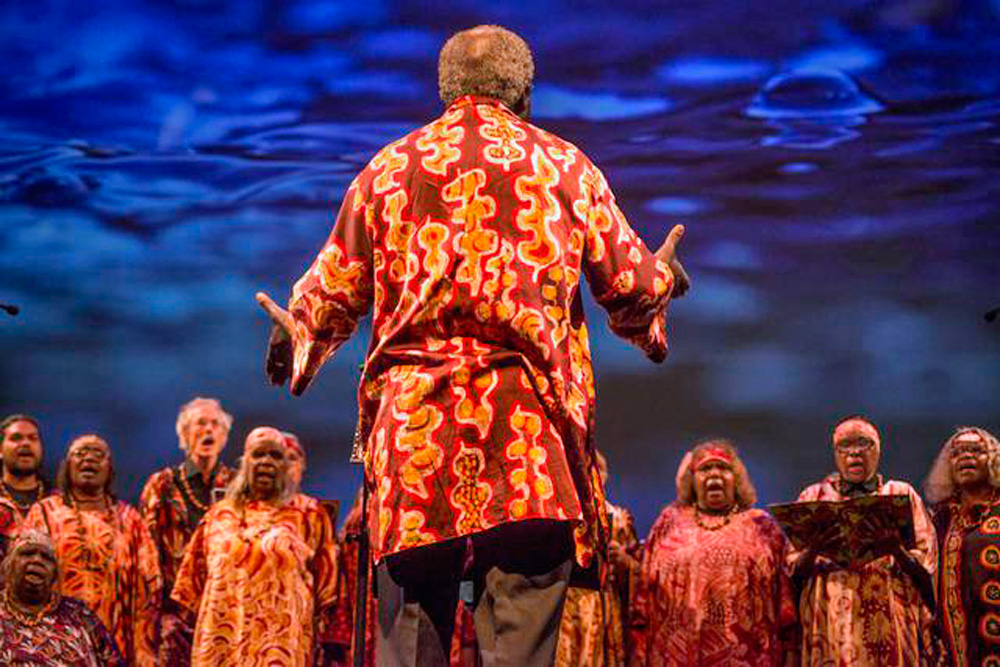 A man with his back to camera standing in front of an Aboriginal choir of around 10 or so women with his arms outstretched, as if conducting them. They are all wearing brightly coloured orange and red clothing.