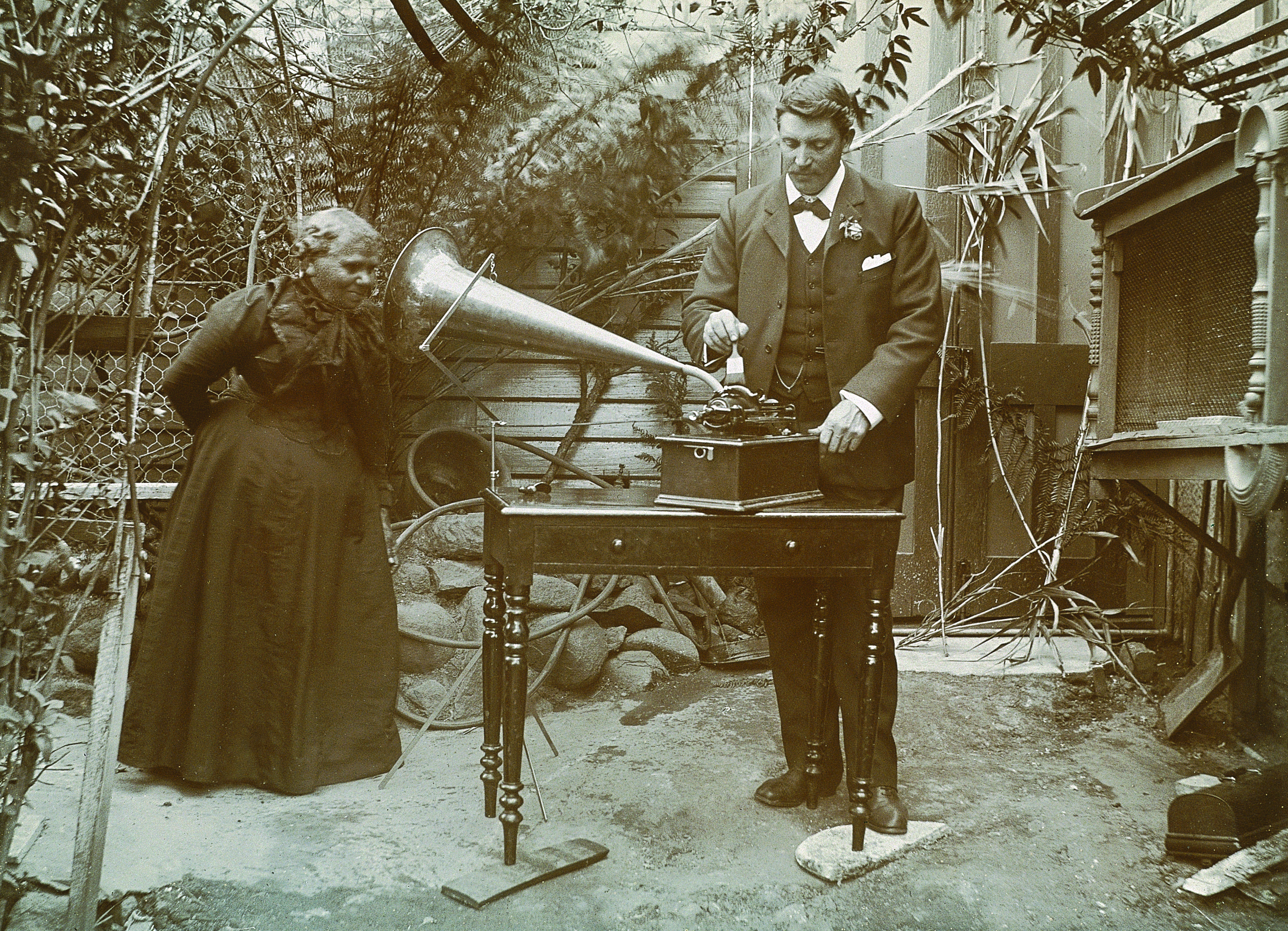 A First Nations woman standing outside and speaking into a gramophone horn while a man stands at a table operating the gramophone