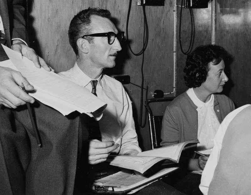 A woman and two men sit in a room. One of the men holds a script in his hand.