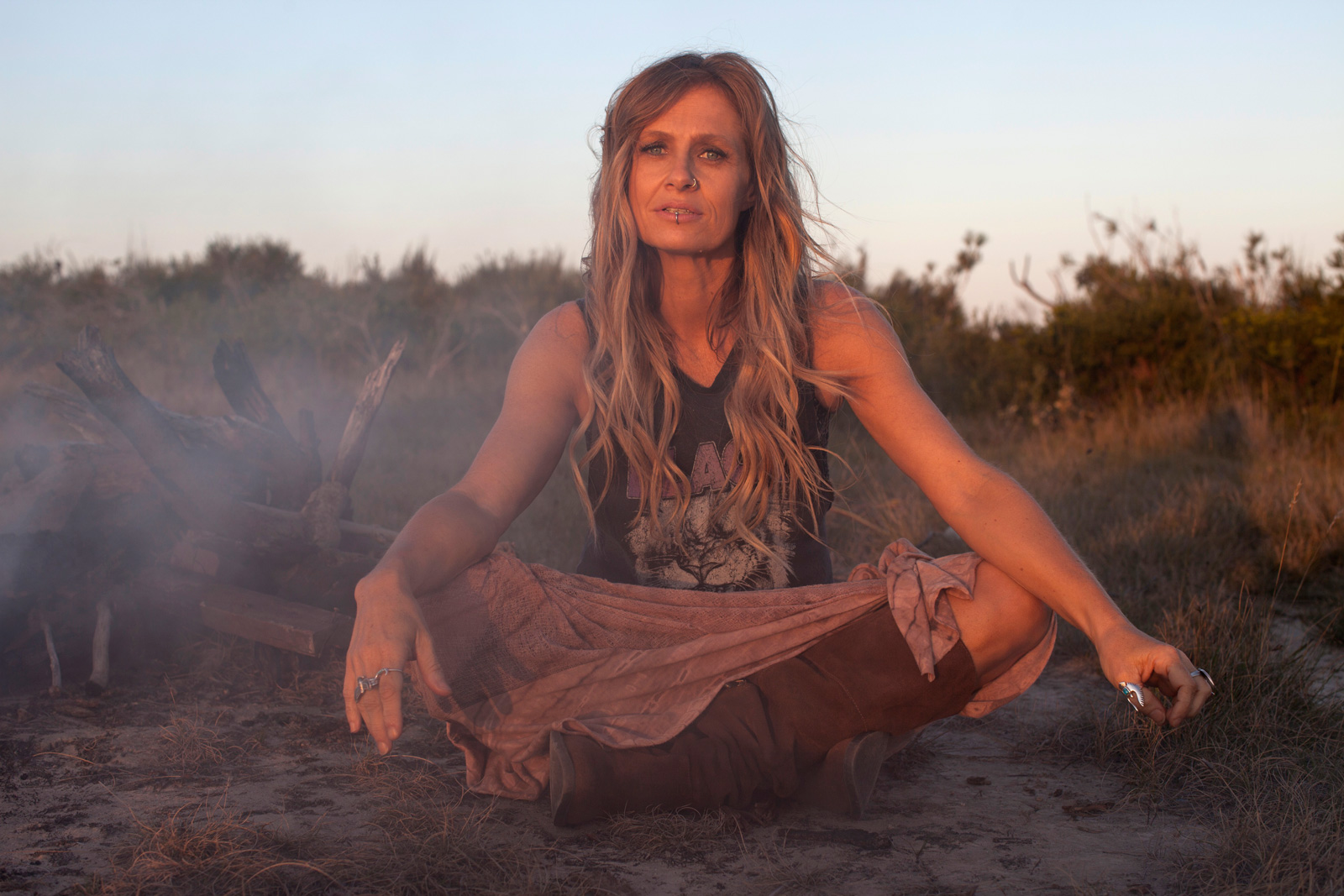 Singer Kasey Chambers sitting cross-legged on the ground in a campfire setting. She is looking directly at camera.