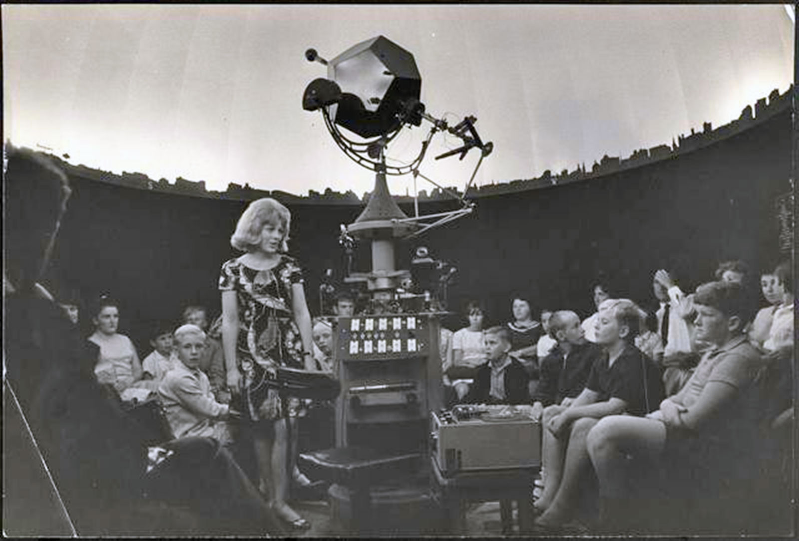 A woman stands in the middle of a crowd of young kids at an observatory