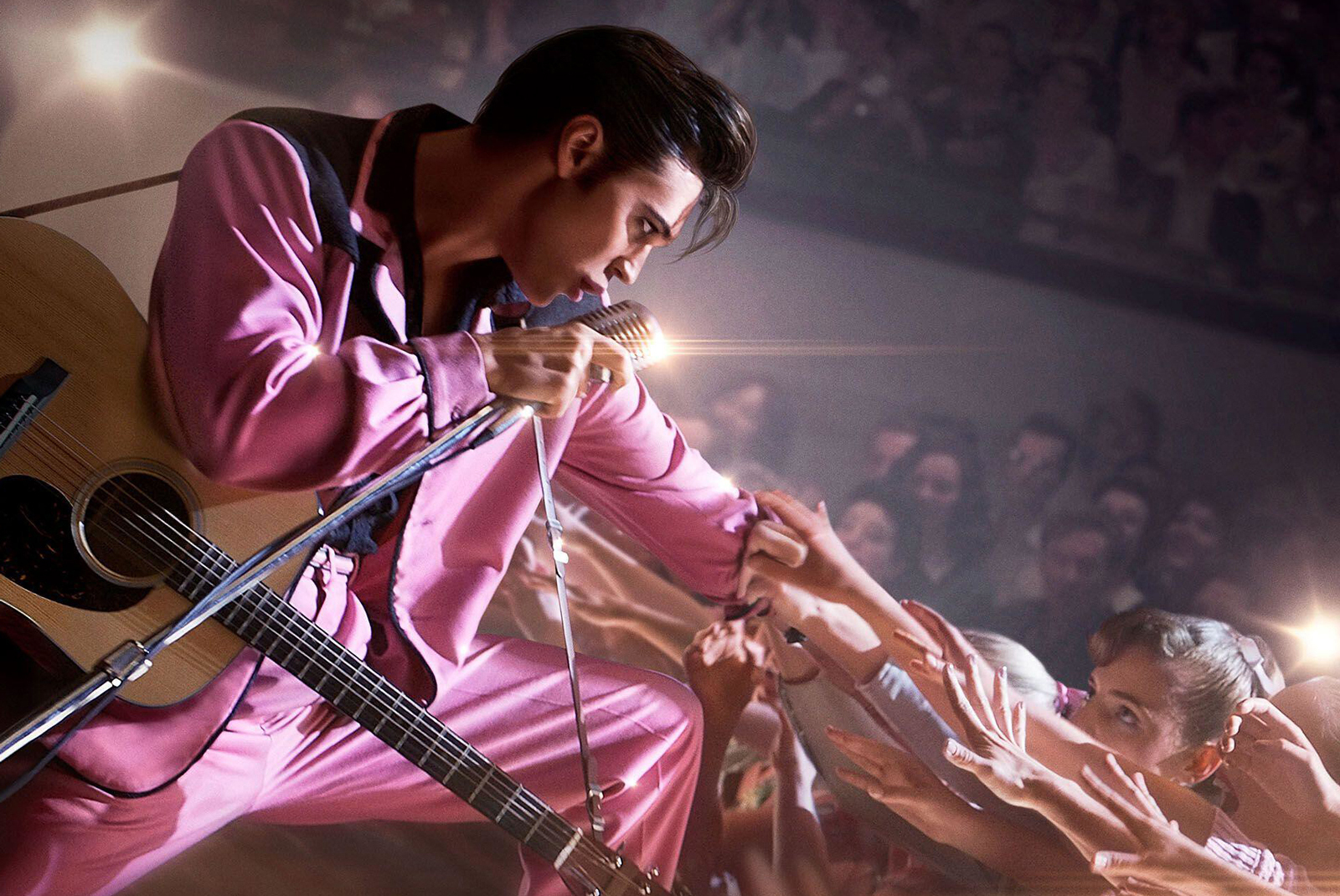 Actor Austin Butler in a scene from the film ELVIS. He is pictured from wearing a bright pink suit and standing on a stage reaching down towards the audience.