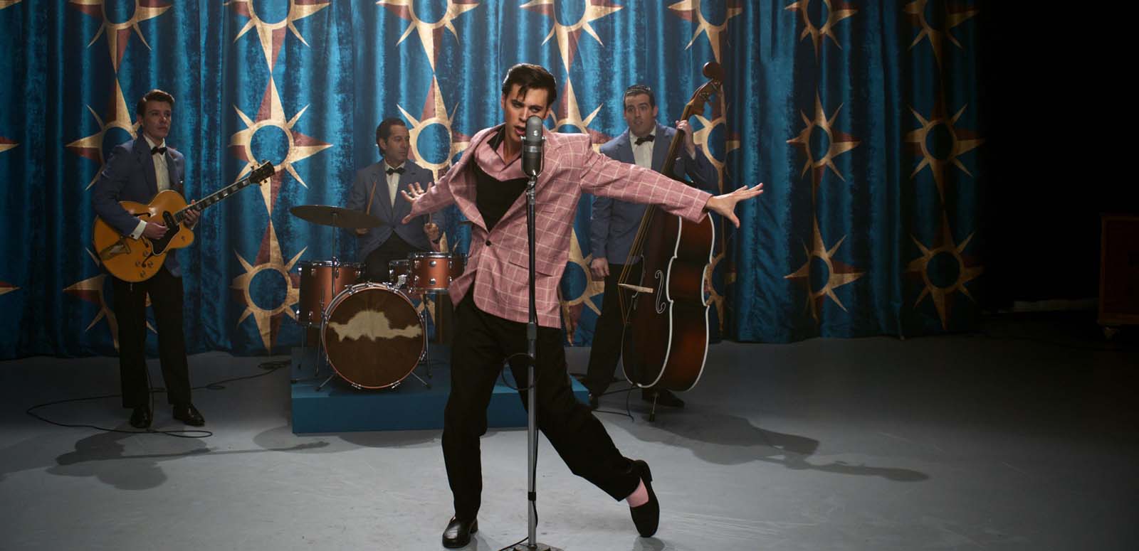 Austin Butler as Elvis Presely singing with his 4-piece band on stage in a recreation of a 1950s TV studio. Elvis is wearing a pink blazer and the group are performing in front of a turqouise curtain with sun symbols on it