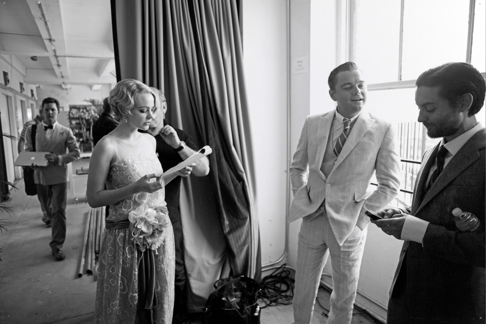 Actors Carey Mulligan, Leonardo DiCaprio and Tobey Maguire standing in a room in a semi-circle. They are on a film set dressed in 1920s costumes.