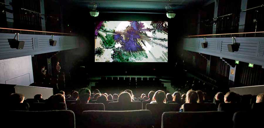 The interior of a cinema pictured from the seats in the back looking towards the screen. There are people in seats watching a film on the screen.