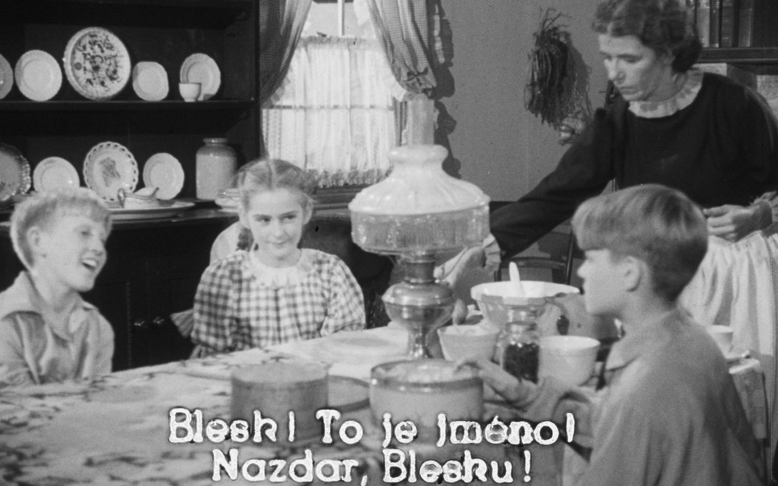 A screenshot from the 1951 film Captain Thunderbolt with Czech subtitles. Three children sit around a table while a woman sets the table