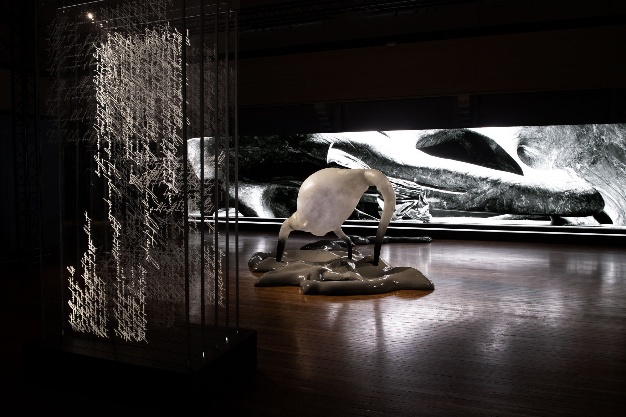 Detail from the art installation Inferno at the NFSA featuring a vertical screen with dense handwriting, a white sculpture arising from a mass on the floor, and a long screen showing a black-and-white landscape.
