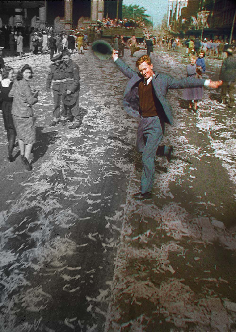 A partly colourised image of a man dancing with raised hat in Sydney, part of a crowd celebrating the end of the Second World War