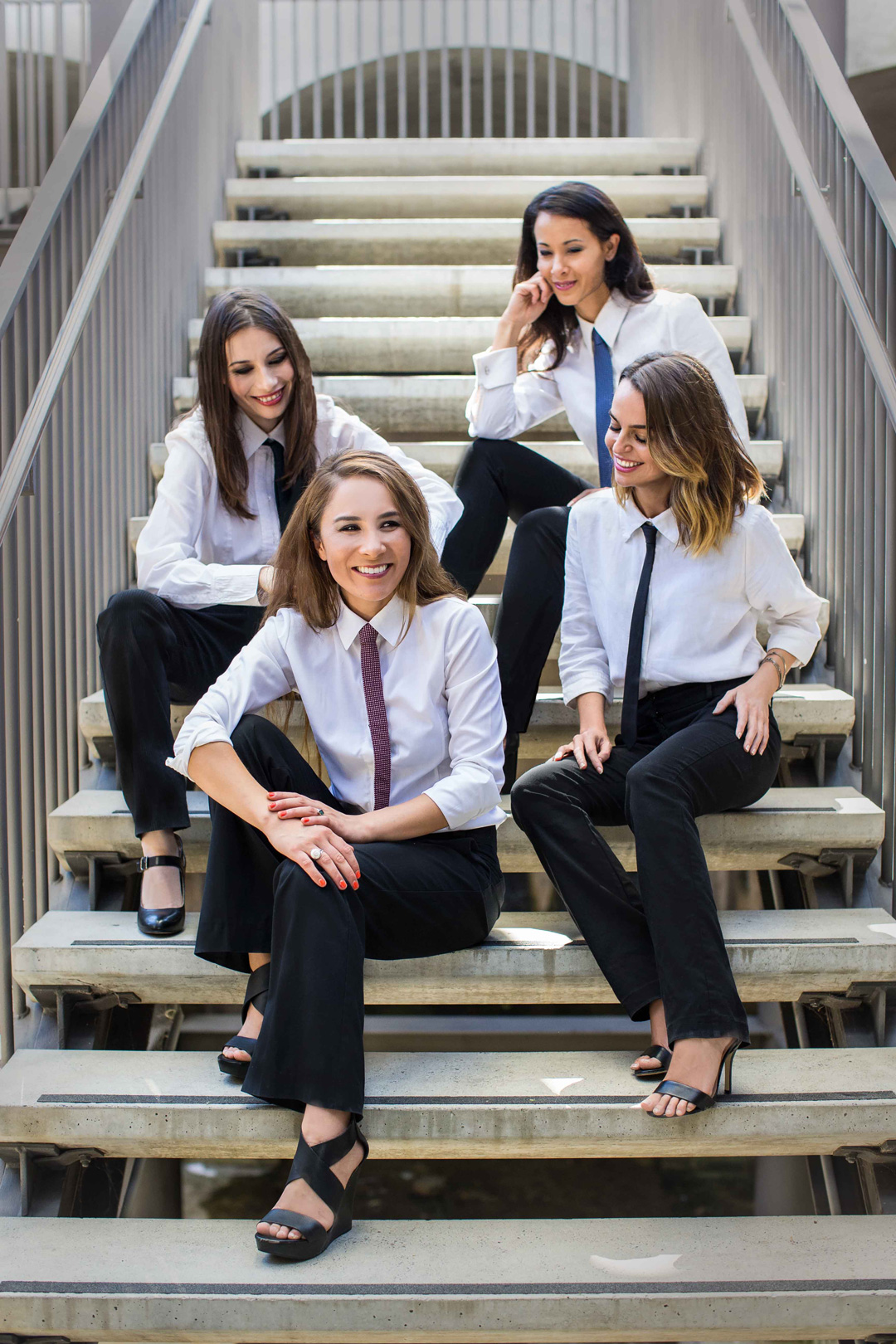 Four young women sitting on a staircase. They are all wearing black pants, white button-down shirts and thin neckties.