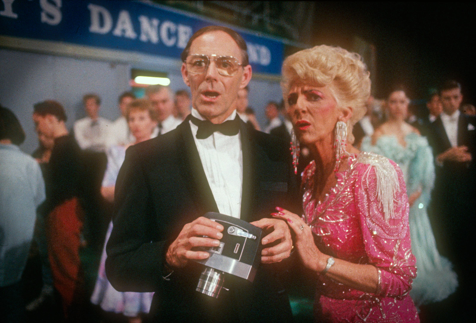 Scene from Strictly Ballroom showing an older man wearing a tuxedo and holding a video camer and an older woman in a pink sequinned dress with a concerned look on their faces. 