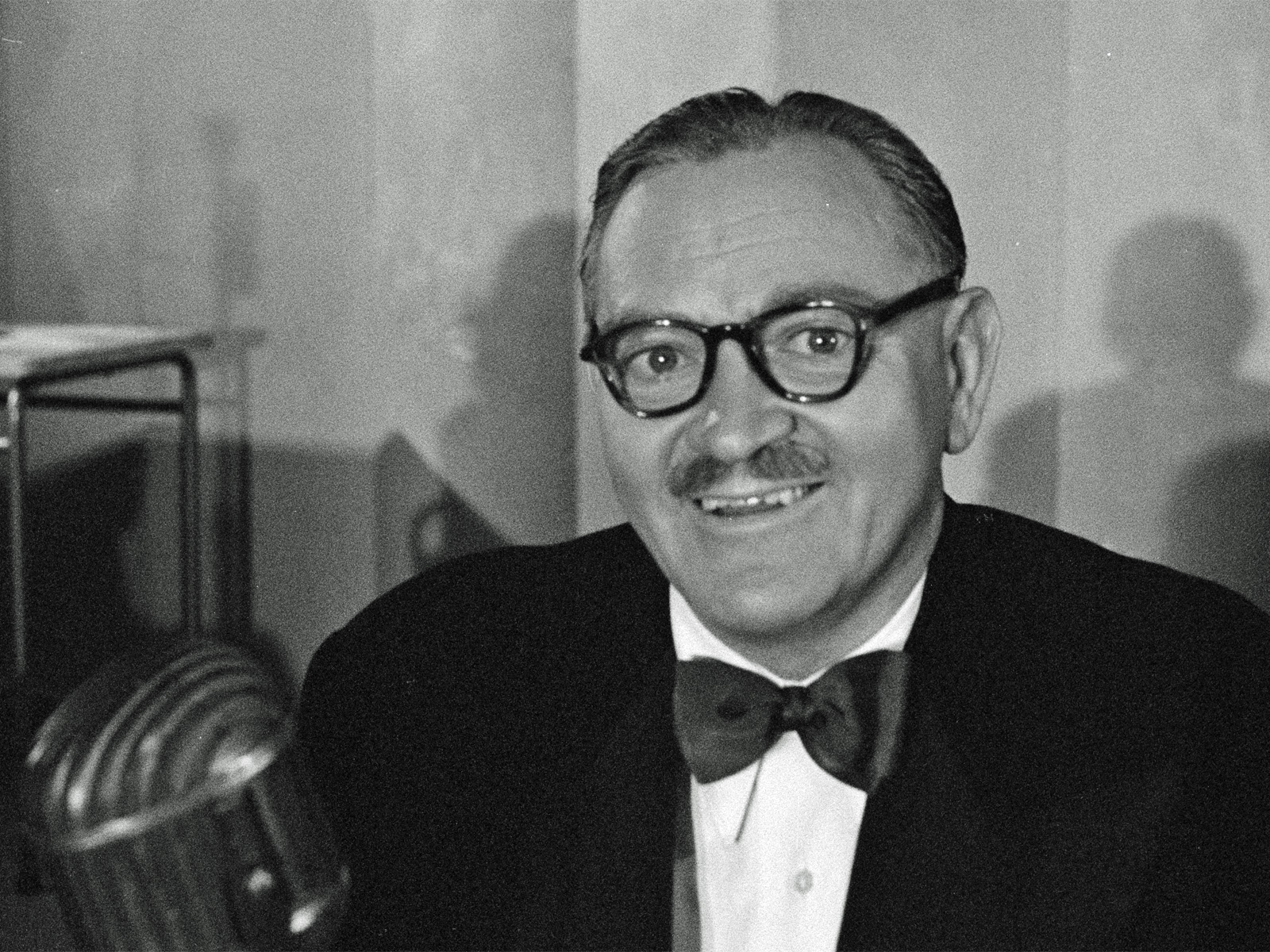 Head and shoulders shot of a man in a suit and bow tie, wearing glasses and smiling at camera.