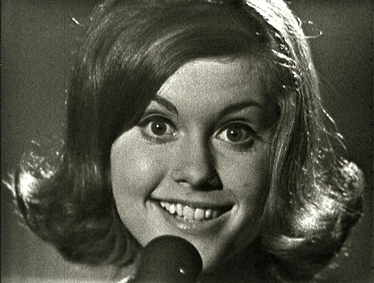 Close up of Olivia Newton-John smiling and looking directly at the camera with a microphone in front of her face, partially covering her chin, from an episode of 