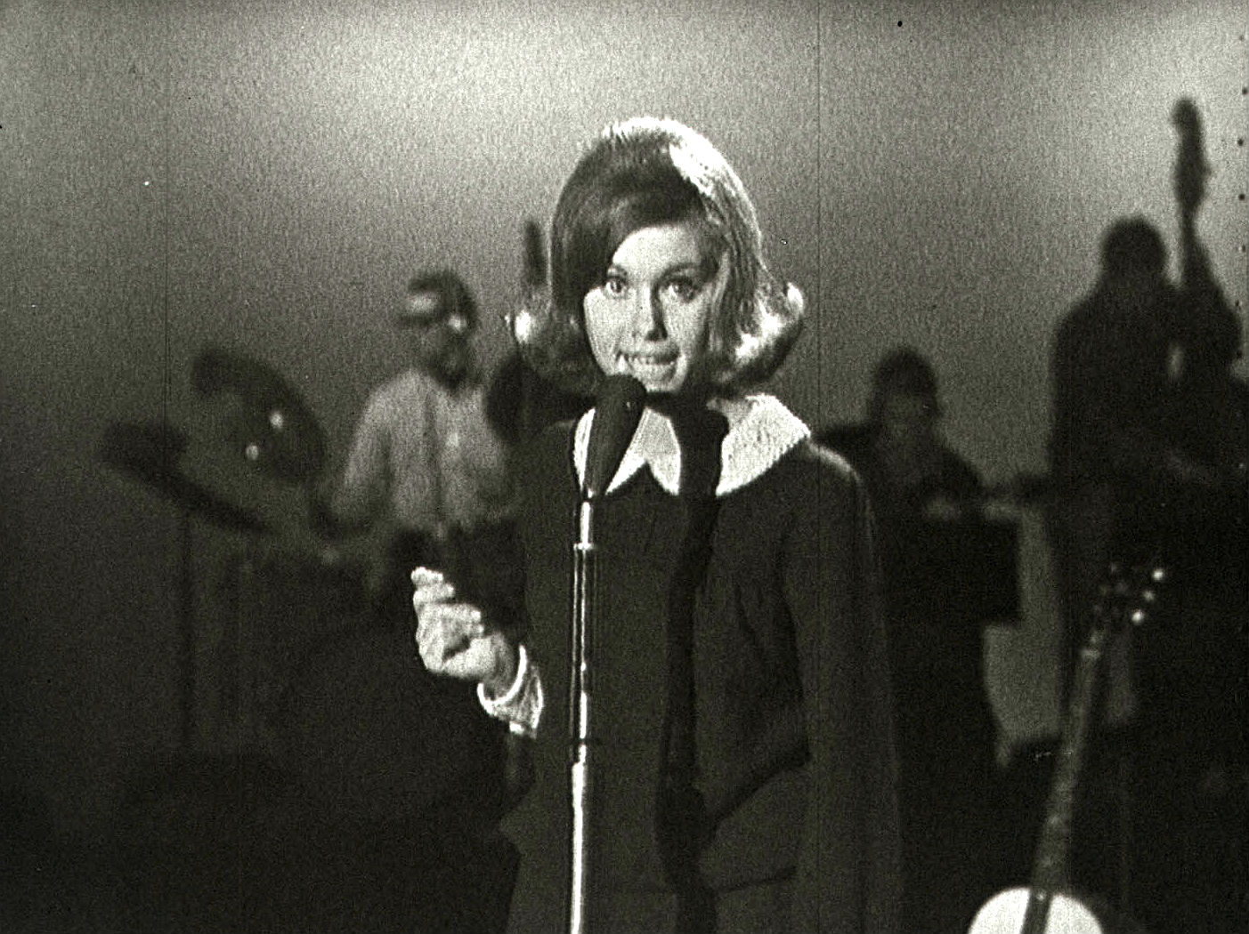 Olivia Newton-John at age 16 in a TV studio standing behind a microphone singing.