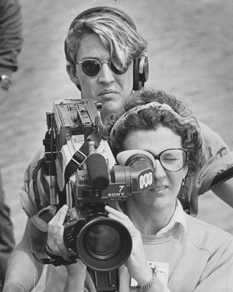 A woman is operating a news camera. A man behind her is wearing headphones.