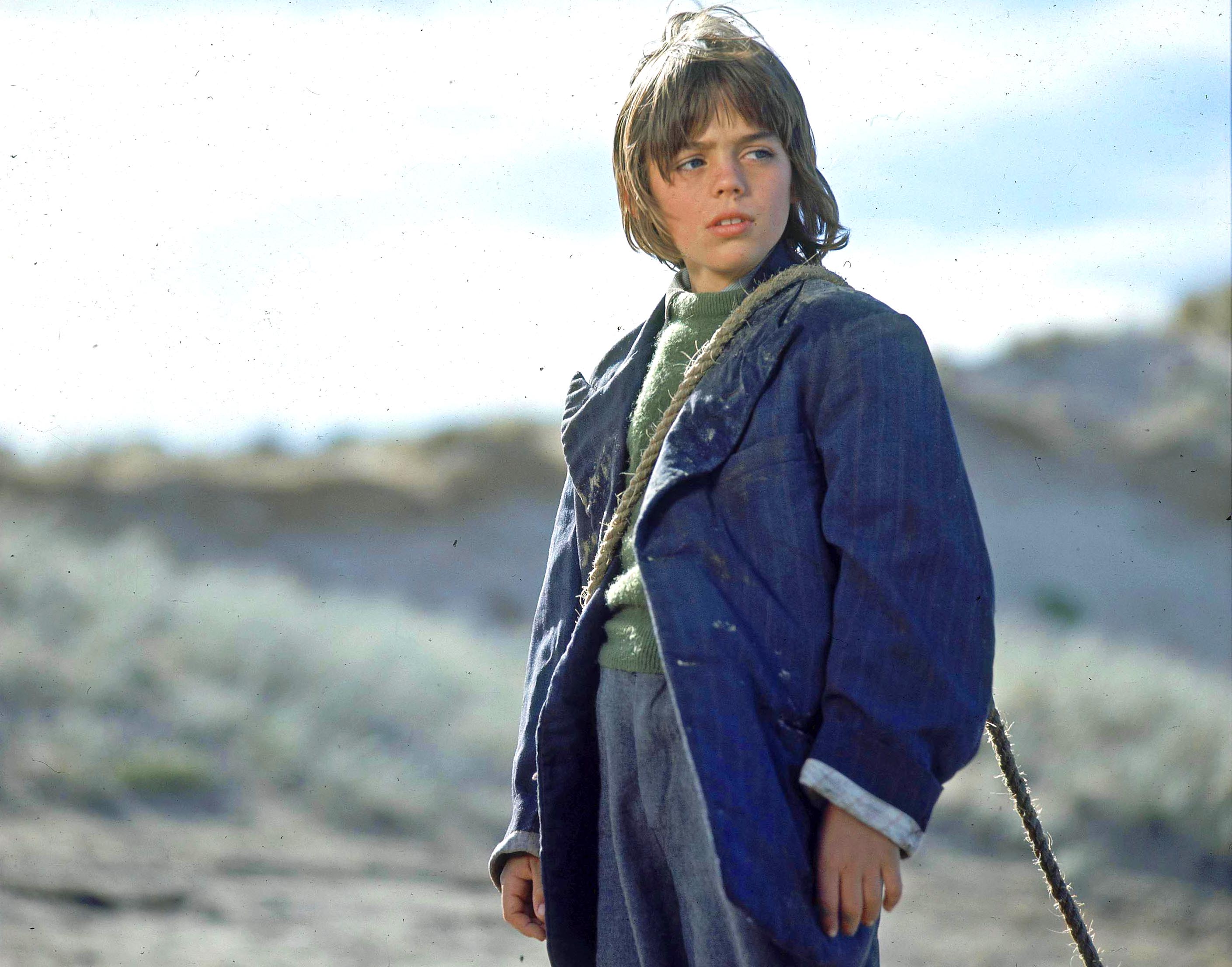 Greg Rowe as Storm Boy, looking deshevelled and dressed in a man's jacket and jumper that are too big for him. He has rope over his shoulder for dragging driftwood back to his humpy