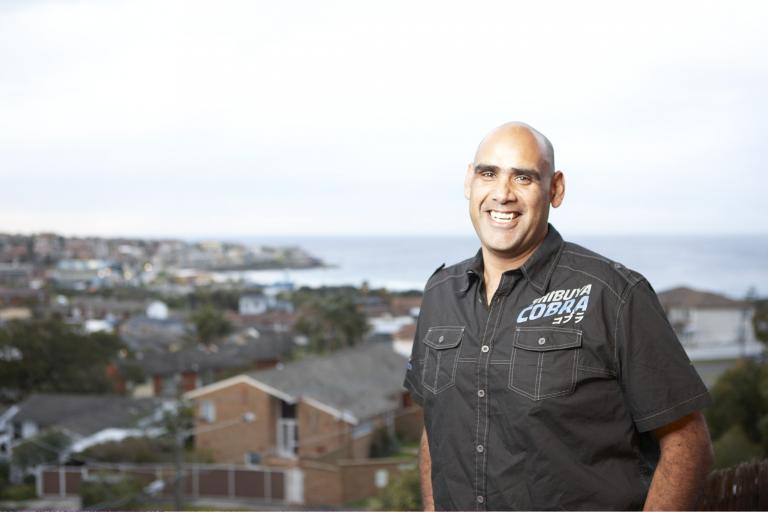 A man in a black shirt smiles in front of a suburban view with the sea in the distance A man in a black shirt smiles in front of a suburban view with the sea in the distance