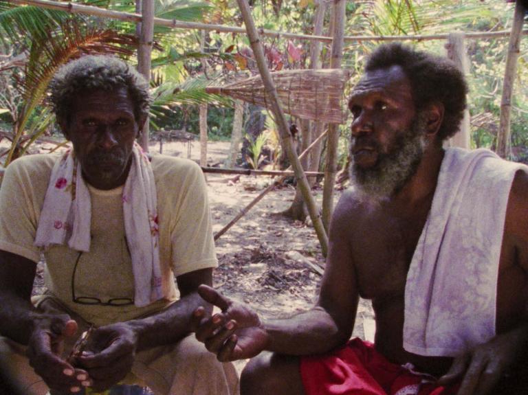 Eddie Mabo is seated on the ground, talking with another man. Eddie Mabo is seated on the ground, talking with another man.