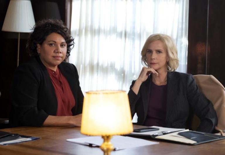 Two women wearing suits seated in a boardroom looking towards camera.