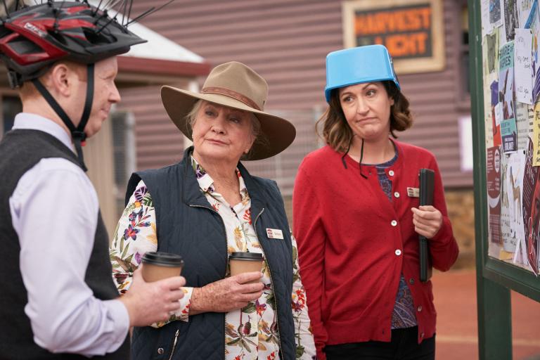 A man and two women all standing together holding coffee cups, the man is wearing a bike helmet and another is wearing an icecream bucket on her head.