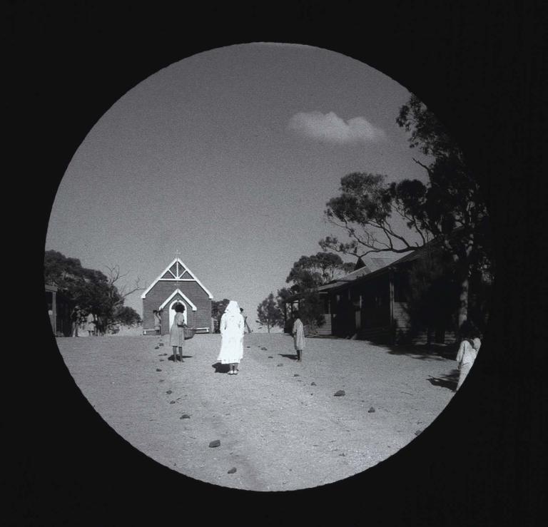 Black-and-white glass slide of a nun with several Indigenous children, as used in Rabbit-Proof Fence. Black-and-white glass slide of a nun with several Indigenous children and a chapel in the background.
