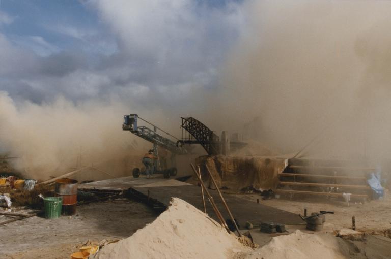 Mad Max Beyond Thunderdome A crew member pushes a crane next to a model set of post-apocalyptic Sydney in some sand dunes. The buildings are about the size of the person. An arch of the destroyed Sydney Harbour Bridge and the Opera House is visible.