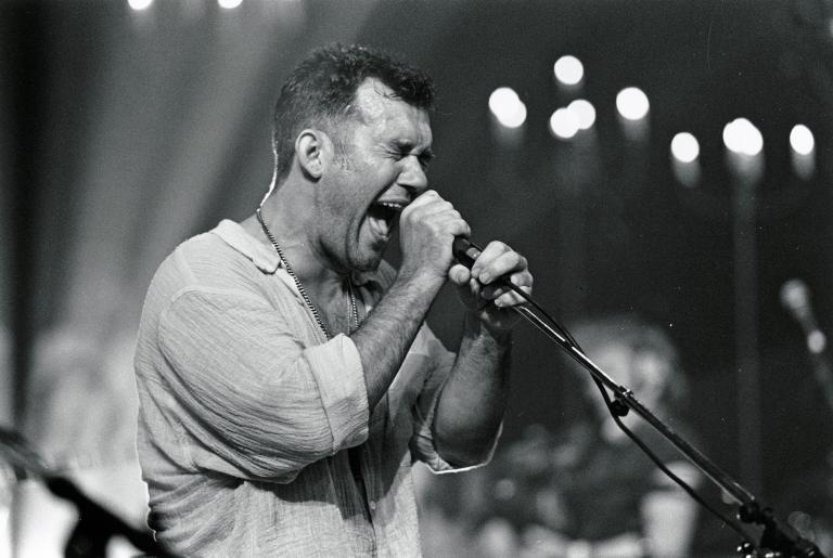 Jimmy Barnes on stage holding a microphone with both hands, singing with eyes closed.