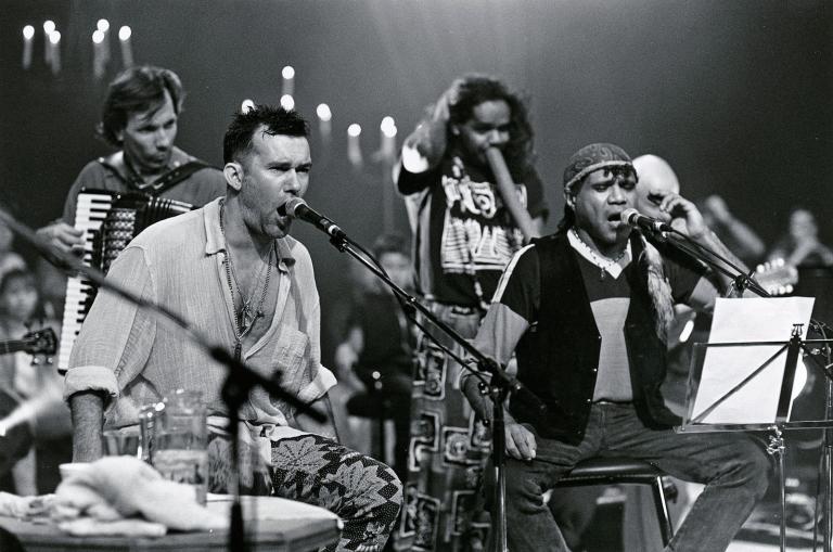 Jimmy Barnes and Archie Roach Singers Jimmy Barnes and Archie Roach on stage with their band during a live concert. Both are seated and singing into microphones.
