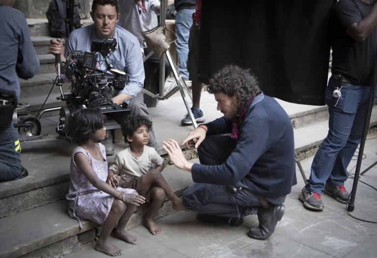 Lion director Garth Davis talking to Sunny Pawar (Young Saroo) and Rita Boy (Amita), who are sitting on a step with cinematographer Greig Fraser setting up a shot behind them