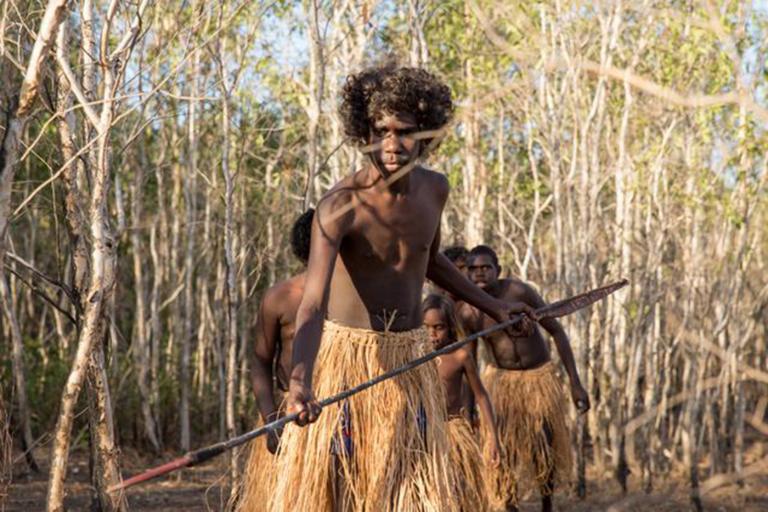 Three Aboriginal youths walking through an area of trees and scrub. They are wearing traditional dress and one is holding a spear.