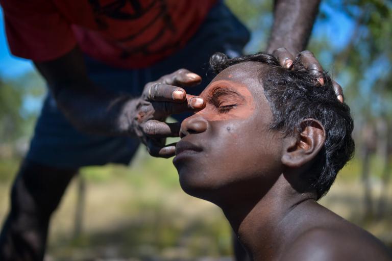 An Aboriginal youth pictured in profile from the shoulders up with his eyes closed is having paint applied to his face by another person. 
