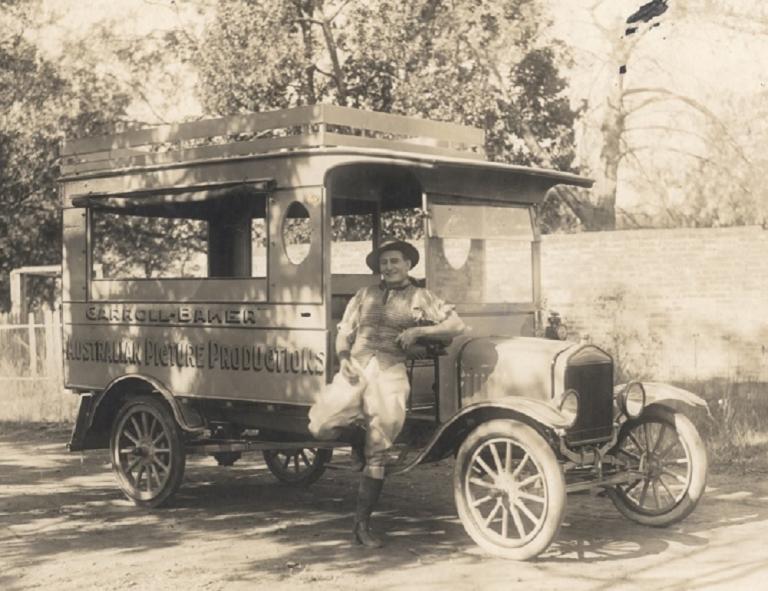 Snowy Baker leans against a truck with 'Carroll-Baker Australian Picture Productions' painted on the side