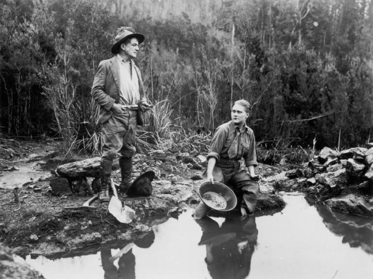 Louise Lovely sits by a stream with a gold pan in a scene from Jewelled Nights.