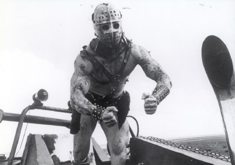 Muscular man wearing an ice hockey mask standing on the back of a truck.
