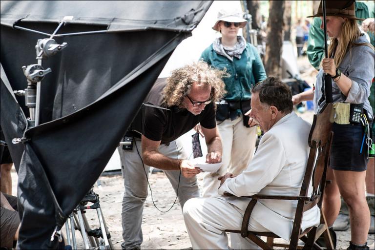 A behind the scenes image from a film production. A man sitting in a wooden chair in a hot, dry outdoor setting. He's wearing a white suit and the director is leaning down speaking to him as some assistants gather around.