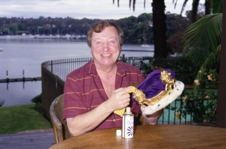 Crown and Brasso Graham Kennedy sitting at a table smiling to camera holding his crown with a bottle of Brasso in front of him