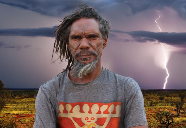 A bearded, middle-aged Aboriginal man in the outback stands staring at camera. In the distant background you can see lighting striking the ground.