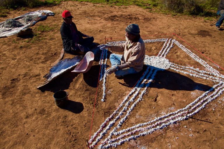 We Don't Need a Map film still Two Aboriginal people sitting in the outback creating an image on the ground using white paint.