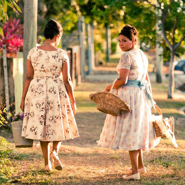 Two women walking away from camera wearing floral dresses, one is carrying a basket and has her head turned back towards the camera.