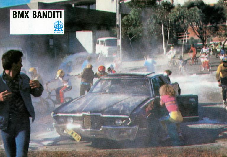 BMX Bandits Yugoslavian Lobby Card A black car with smoke coming out of it and the front licence plate falling off. The car is surrounded by bystanders and one girl who is kneeling down and looking into one of the car doors.
