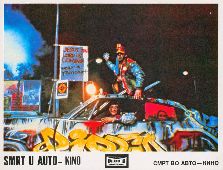 Dead End Drive-In Yugoslavian Lobby Card A man driving a beaten up, graffitied car with another man riding on top. Both men look menacing and threatening, as if about to attack someone. In the background is more graffiti on dilapidated fence.