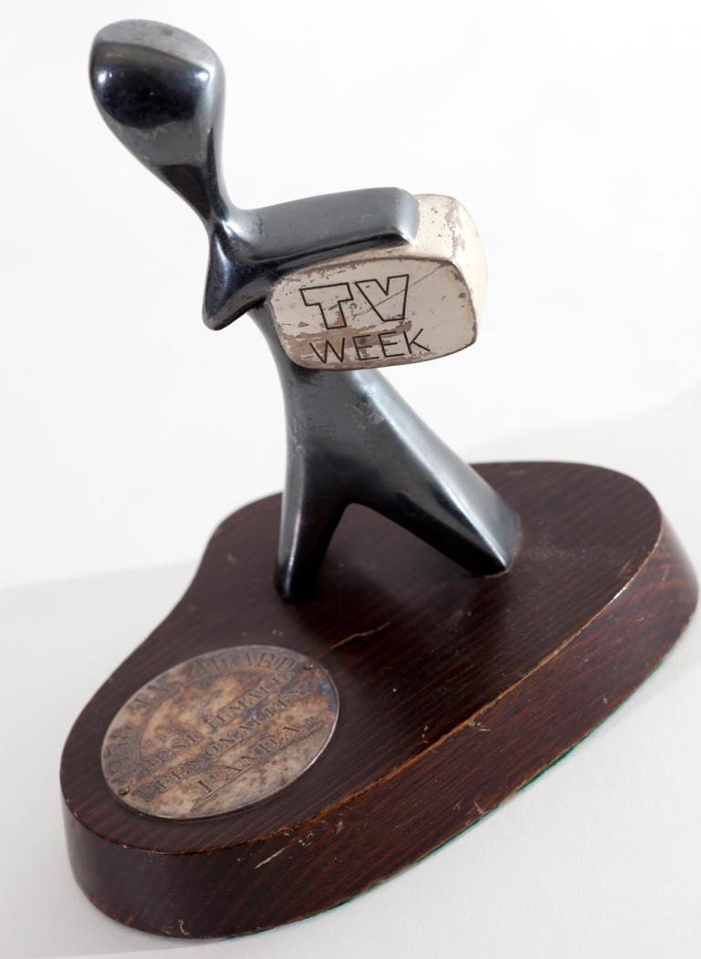 A TV Week Logie Award, made up of a small silver statuette in the shape of a man holding a television. The statuette sits on a round wooden base with a metal disc with words engraved on it. 
