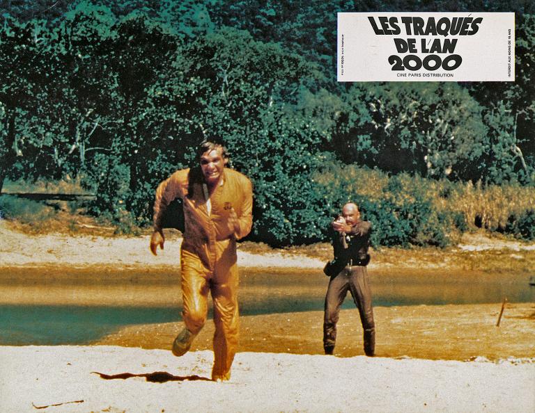 Turkey Shoot French Lobby Card - 'Paul Anders' A prisoner running away from another man pointing a gun on the bank of a river in a scene from the film Turkey Shoot.