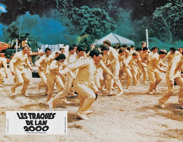A large group of men and women in yellow prison uniforms running away from their captors in a scene from the film Turkey Shoot.