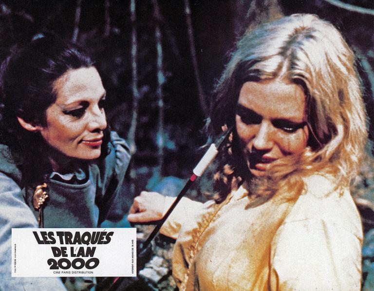Turkey Shoot French Lobby Card - 'Jennifer and Rita' Close up of a menacing-looking dark-haired woman (Carmen Duncan) tormenting a blond woman (Lynda Stoner) by pointing an arrow at her face in a scene from 'Turkey Shoot'.