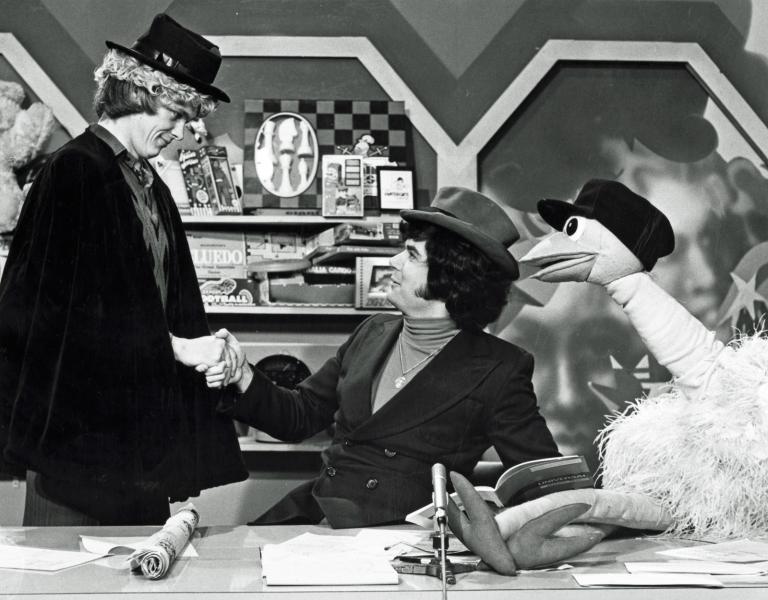 John Farnham dressed in hat and cape, shaking hands with Daryl Somers, seating at a desk, also wearing a hat and Ossie Ostrich seated beside him wearing a cap.