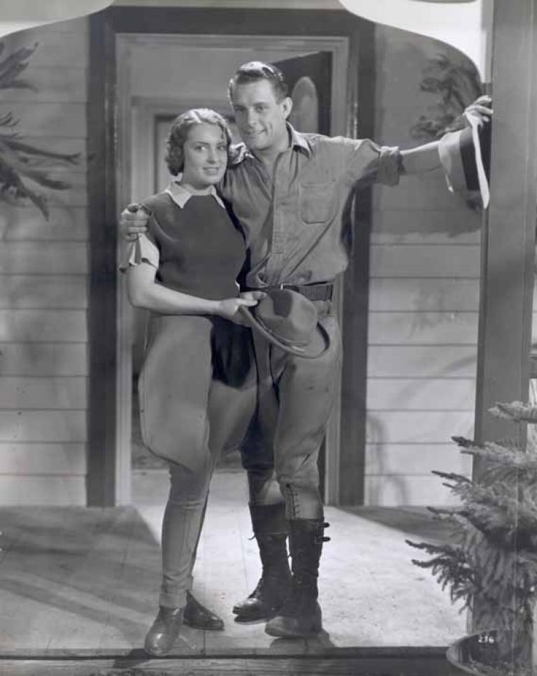 Full length image of a young woman and man standing on a front porch of a house. The man has his arm around the woman's shoulder. They are dressed in riding attire.