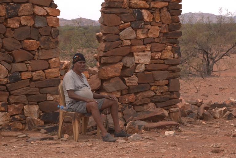 Jupurrurla – Man of Media A man sits on a chair in front of a stone wall on a plain in the outback.